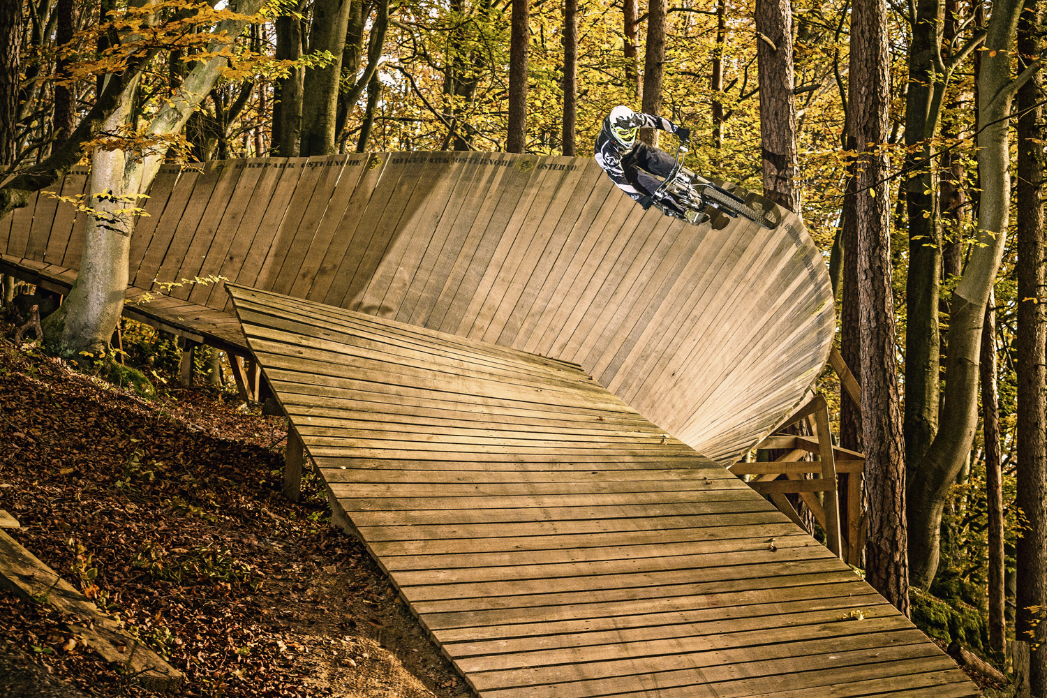 Ein Radfahrer, fotografiert mit der Präzision eines kommerziellen Sportfotografen, trägt Schutzkleidung, während er gekonnt um eine Holzwand in einem dichten Wald navigiert.