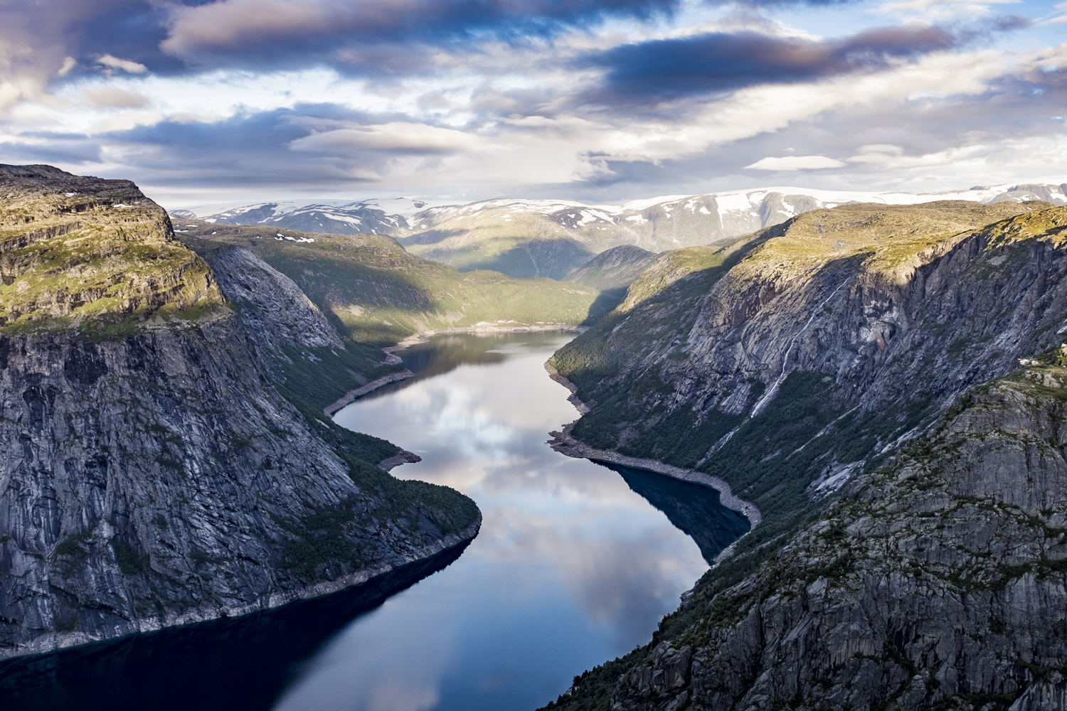 Ein ruhiger Fjord, in dessen stillem Wasser sich der Himmel spiegelt, umgeben von steilen Felsklippen und in der Ferne schneebedeckten Bergen unter einem teilweise bewölkten Himmel – eine ideale Kulisse für kommerzielle Fotografie, die die Erhabenheit der Natur einfängt.