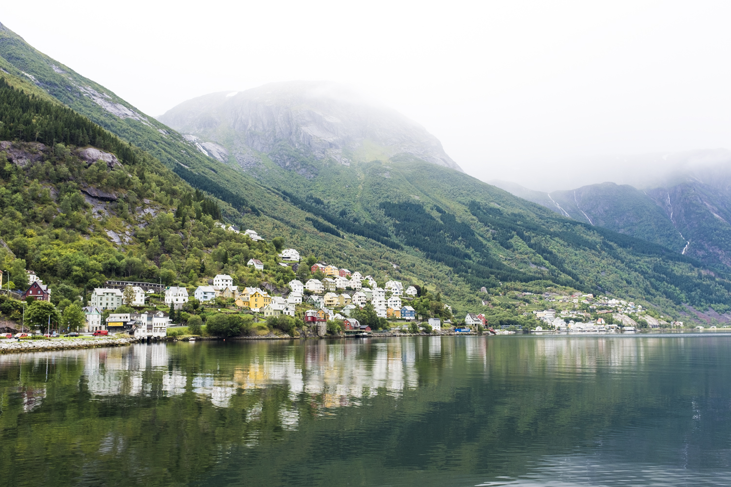 Malerische Aussicht auf ein malerisches Dorf mit farbenfrohen Häusern, eingebettet in einen ruhigen Fjord, ideal für ein kommerzielles Shooting, umgeben von üppig grünen Bergen unter einem bewölkten Himmel.