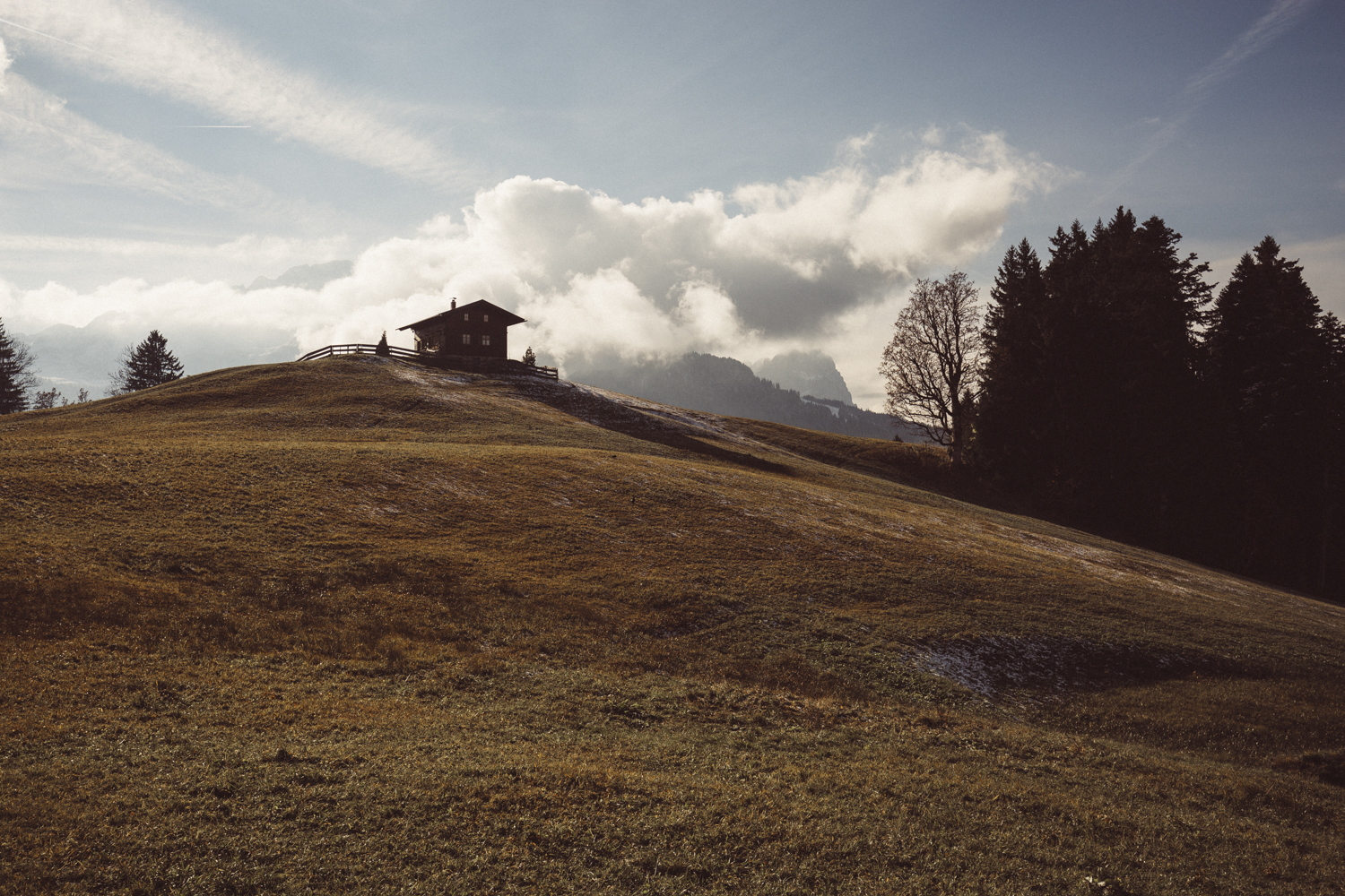 Ein kleines Holzhaus steht auf einem grasbewachsenen Hügel mit Bäumen im Hintergrund und einem bewölkten Himmel und bietet die perfekte Kulisse für Business-Fotografie, die die Essenz des rustikalen Charmes in kommerziellen Aufnahmen einfängt.