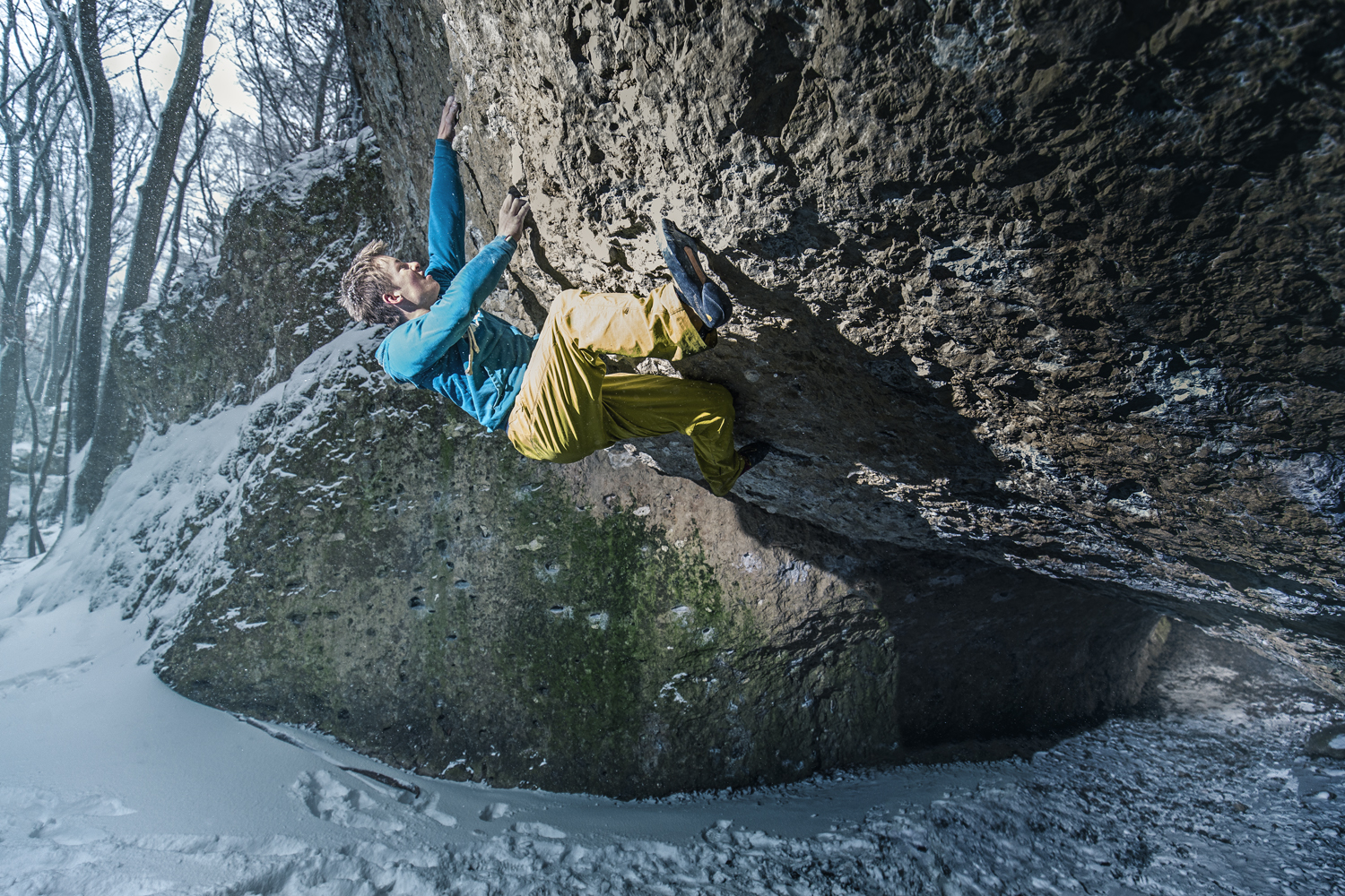 Eine Person mit einer blauen Jacke und gelben Hosen klettert auf einen großen Felsüberhang in einem verschneiten Wald und fängt in dieser Werbeaufnahme des Werbefotografen Nürnberg perfekt den Geist des Abenteuers ein.