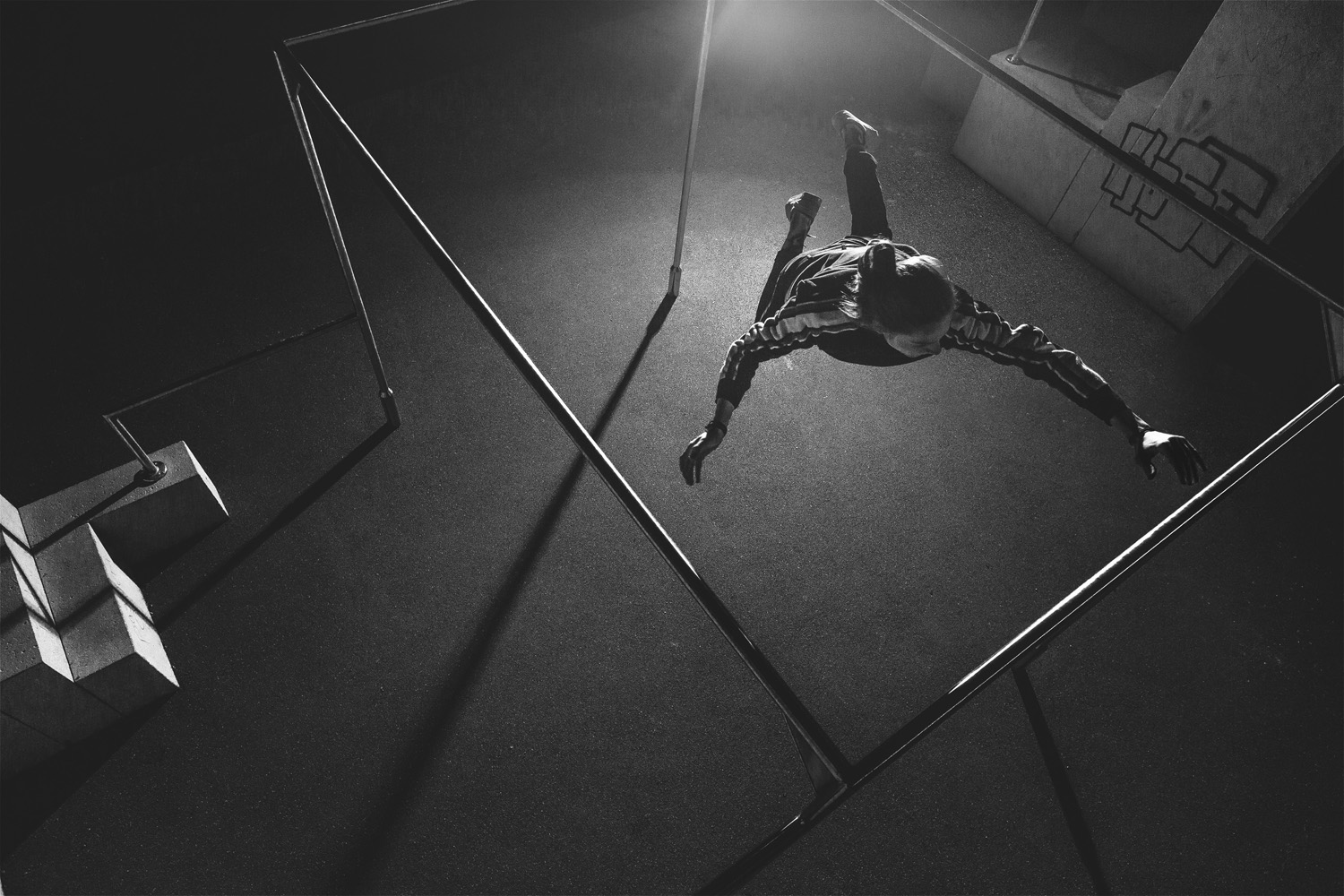 Eine Person, die in der Luft einen Parkour-Sprung über ein Geländer in einer städtischen Nachtkulisse ausführt, von einem Sportfotografen aus der Luft in Schwarzweiß aufgenommen, zeigt die dynamische Bewegung und urbane Ästhetik, die problemlos das Portfolio jedes Fotostudios zieren würde.