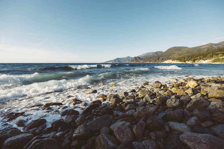 Eine felsige Küste mit brechenden Wellen unter einem klaren blauen Himmel schafft eine ruhige Kulisse für kommerzielle Fotografie. In der Ferne säumen Hügel den Horizont und bieten eine ideale Kulisse, um die Essenz der Businessfotografie in der Ruhe der Natur einzufangen.