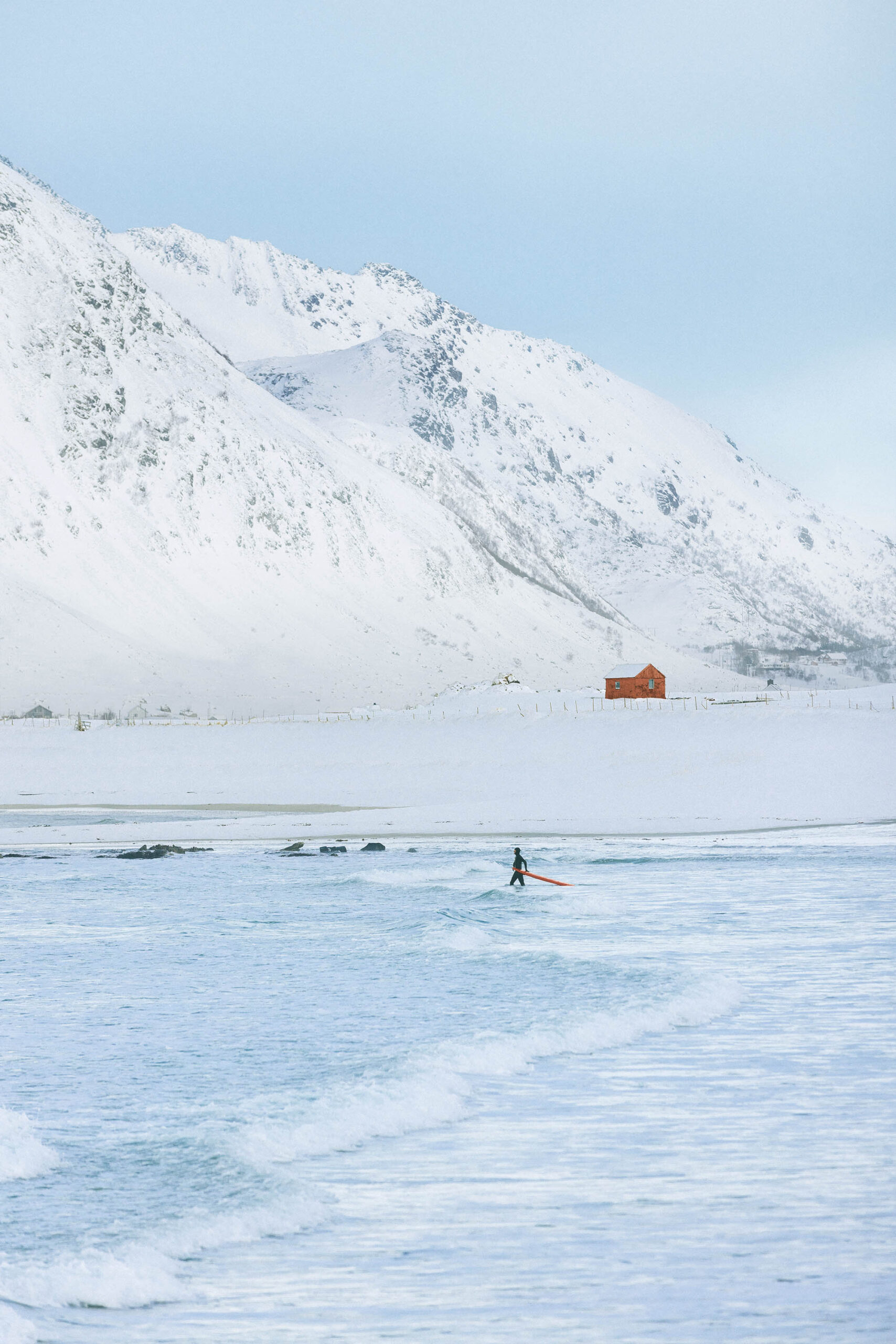 Eine Person surft im Meer in der Nähe einer schneebedeckten Küste, eingerahmt von majestätischen Bergen und einem einzelnen roten Gebäude, und verkörpert den Geist eines kommerziellen Abenteuers.