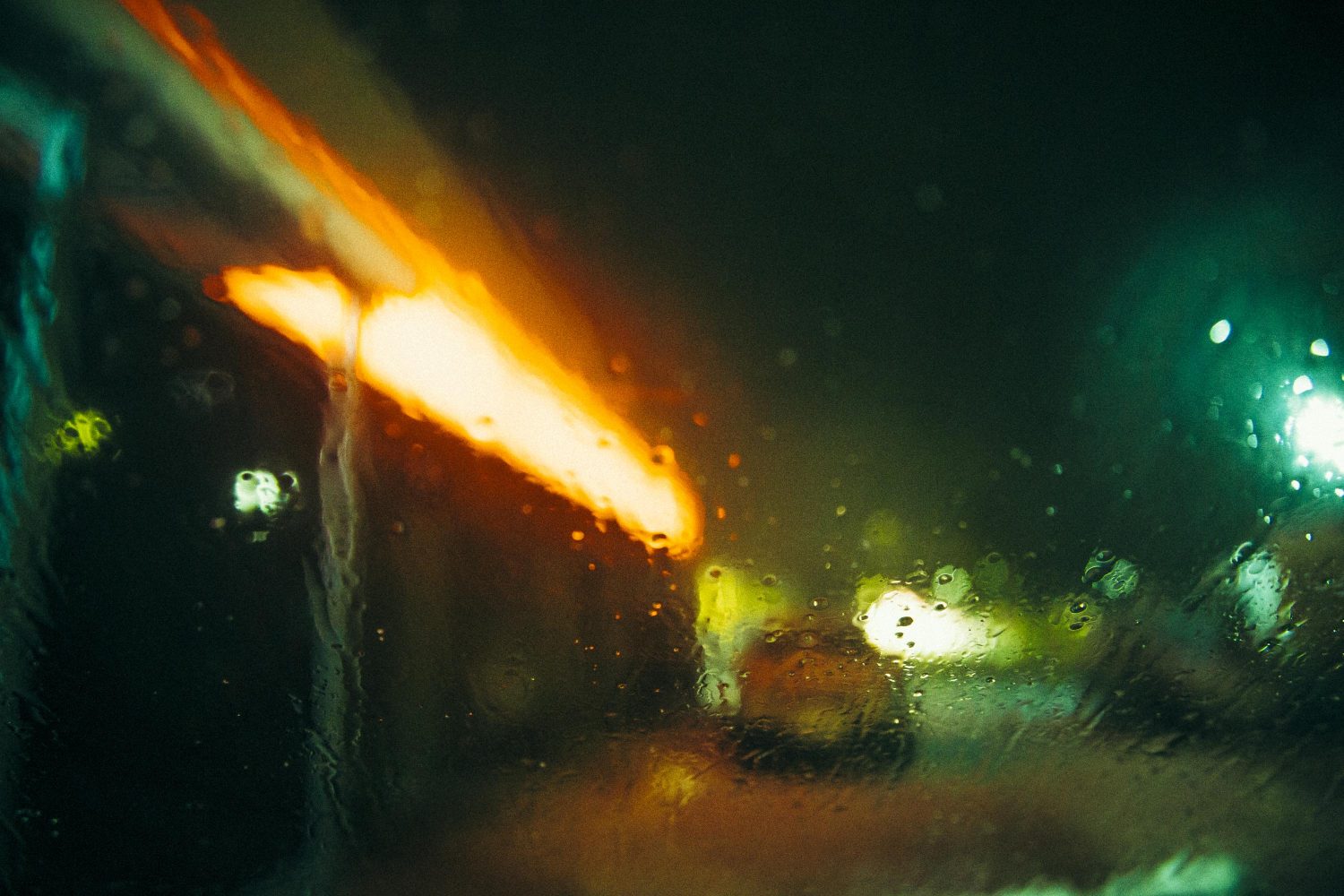 Abstrakter Blick durch ein regennasses Fenster bei Nacht, der an eine stimmungsvolle Fotosession im Business-Studio erinnert, mit verschwommenen orangefarbenen Straßenlaternen und subtilen grünlichen Farbtönen.